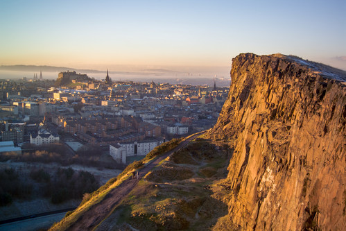 A pointed crag of rock with a footpath at the base rises in front of a cityscape, with a castle on a hill, in the background